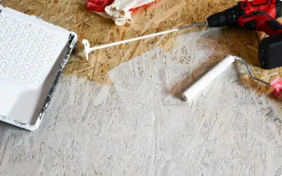 Beim Streichen von OSB-Platten mit weißer Farbe: Ein Farbroller und eine Farbwanne liegen bereit. Handschuhe und ein Akkuschrauber sind ebenfalls zu sehen.