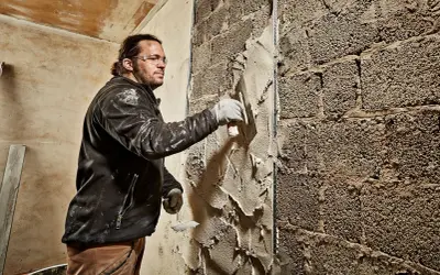 Mann verputzt eine grobe Ziegelwand mit einer Kelle. Der Putz wird gleichmäßig aufgetragen, um die Oberfläche zu glätten.