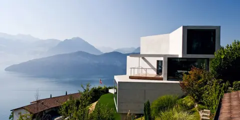 Modernes Haus in Hanglage mit Blick auf einen See und Berge. Ein traditionelles Haus liegt tiefer am Hang.