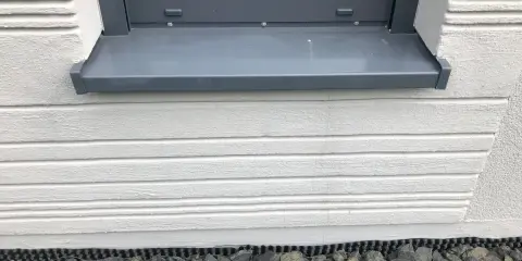 Graue Fensterbank mit leichter Neigung, die Regenwasser ableitet. Darunter weiße Holzverkleidung und grauer Schotter.