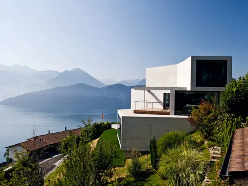 Modernes Haus in Hanglage mit Blick auf einen See und Berge. Ein traditionelles Haus liegt tiefer am Hang.