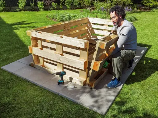 Mann baut ein **Hochbeet aus Paletten** im Garten. Werkzeug und Material liegen bereit.