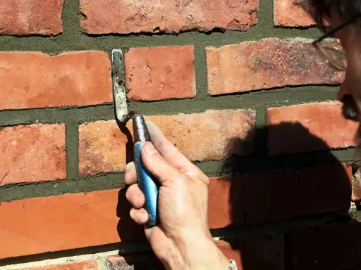 Hand mit Kelle bessert Zementfugen einer Ziegelwand aus.