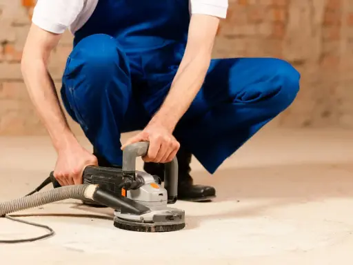 Mann in blauer Latzhose schleift mit einer Maschine den Estrich. Staub wird abgesaugt.