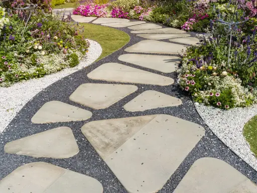 Ein geschwungener Weg aus unregelmäßigen Trittsteinen im Garten, umgeben von bunten Blumen und Kies. Perfekt, um Trittsteine Garten selber machen.
