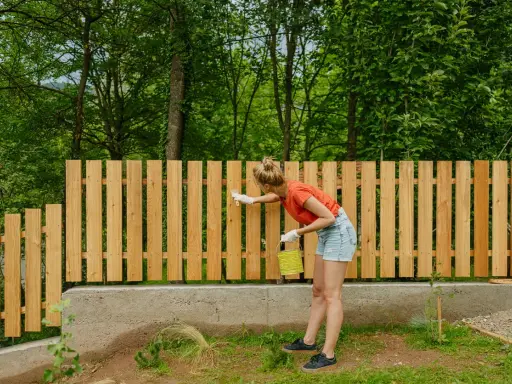 Frau streicht neuen Holzzaun, um ihn vor Witterungseinflüssen zu schützen. Sie setzt den Zaun mit Sorgfalt und Hingabe.