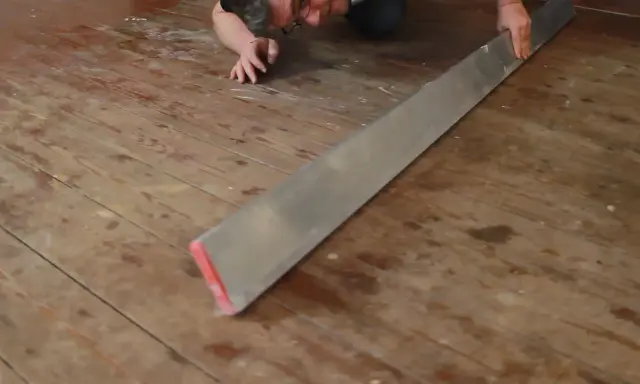 Mann verlegt Dielenboden, hält ein langes Metallwerkzeug, um die Bretter auszurichten.
