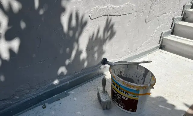Werkzeug und Farbeimer bereit zum Ausbessern des Sockelputzes. Schatten von Blättern fallen auf die Wand.