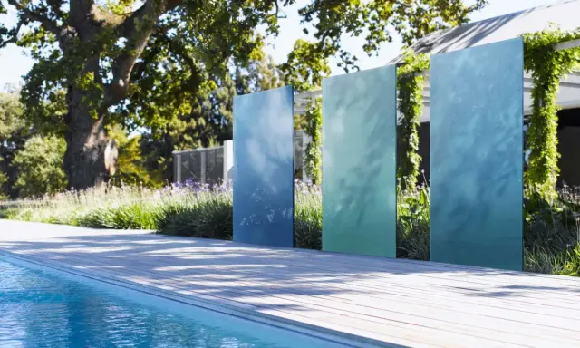 Ausgefallener Sichtschutz Garten selber machen: Vertikale, blaue und grüne Metallplatten vor üppiger Bepflanzung und Pool.