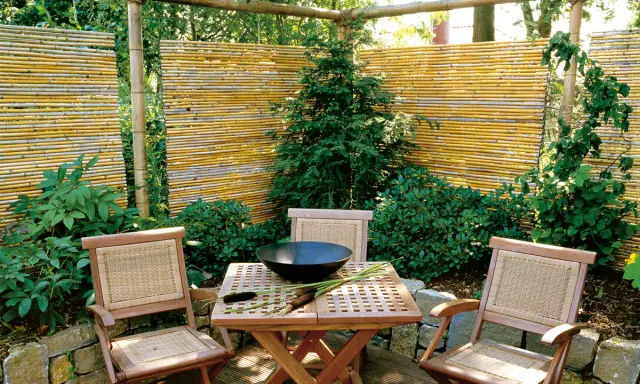 Ein lauschiger Sitzplatz mit einem selbstgemachten Zaun aus Bambus. Holzmöbel und üppige Pflanzen schaffen eine natürliche Atmosphäre.