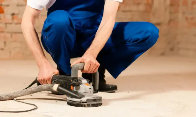 Mann in blauer Latzhose schleift mit einer Maschine den Estrich. Staub wird abgesaugt.