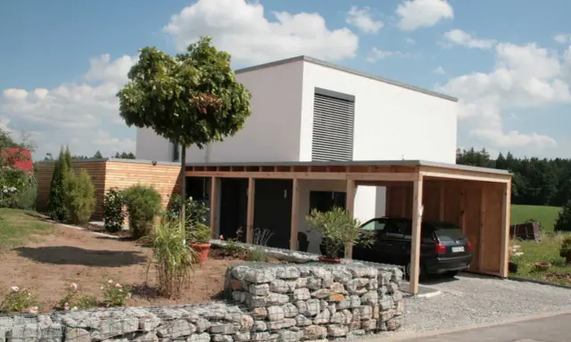 Modernes, weißes Haus mit Holzcarport und Gabionenmauer. Ein Beispiel, wie man ein low budget haus bauen kann, das dennoch stilvoll aussieht.