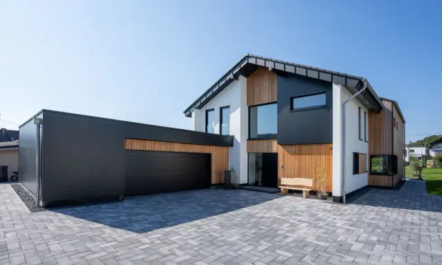 Modernes Haus mit Garage, dessen Satteldach modern interpretiert ist. Holz und dunkle Fassadenelemente prägen das Bild.