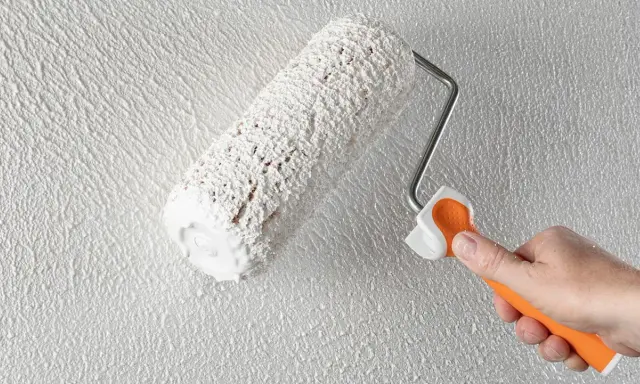 Eine Hand mit orangefarbenem Griff hält eine Farbrolle, die weißen Putz auf eine strukturierte Wand aufträgt. Der Anschluss Putz an Holz wird hier perfektioniert.
