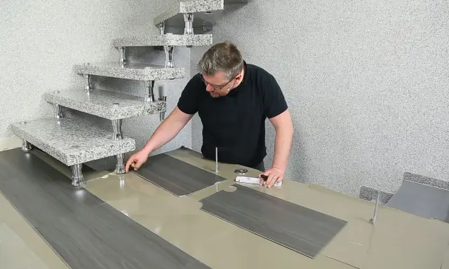 Mann verlegt PVC-Boden. Die grauen Planken werden passgenau zugeschnitten und neben einer modernen Treppe ausgelegt.
