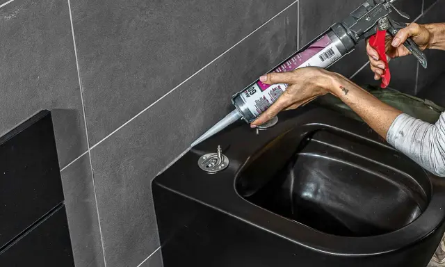Person erneuert silikonfugen am schwarzen Bidet mit einer Kartuschenpistole.