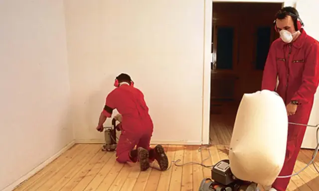 Zwei Männer in roten Overalls schleifen Dielen ab. Einer kniet und bedient eine Schleifmaschine, der andere steht und hält einen Staubfangsack.