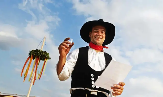 Ein Mann im Zimmermannsgewand hält ein Glas und eine Urkunde. Ein Richtkranz hängt am Himmel. Das ist ein Richtfest!