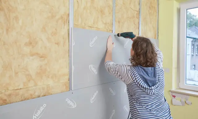 Frau verkleidet Wand mit Rigipsplatten und Schrauber. Isolierung ist sichtbar.