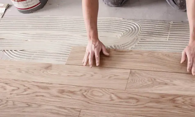 Hände legen Holzplanken auf Kleber, um den Boden zu verlegen.