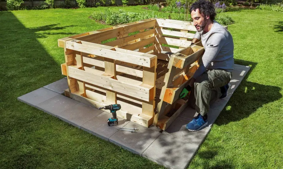 Mann baut ein **Hochbeet aus Paletten** im Garten. Werkzeug und Material liegen bereit.