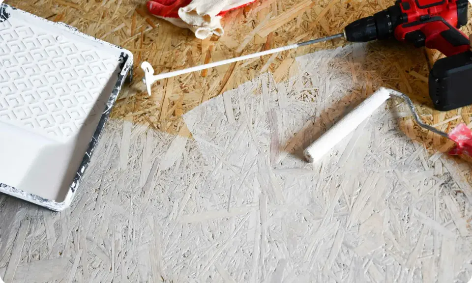 Beim Streichen von OSB-Platten mit weißer Farbe: Ein Farbroller und eine Farbwanne liegen bereit. Handschuhe und ein Akkuschrauber sind ebenfalls zu sehen.