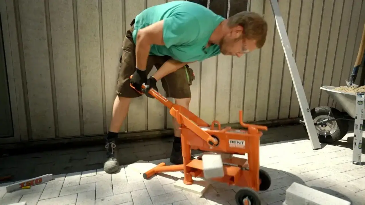 Mann mit Schutzbrille und Handschuhen nutzt eine orangefarbene Maschine, um Terrassenplatten zu schneiden. Werkzeuge und Schubkarre im Hintergrund.