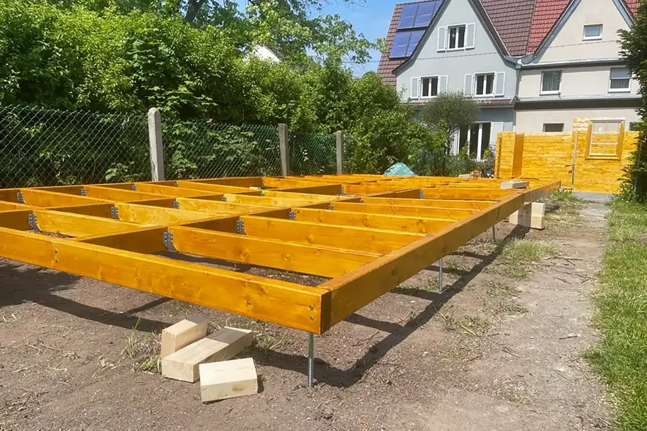 Holzbalken für den Gartenhaus Boden werden auf Pfosten montiert. Ein Teil des Gartenhauses steht bereits im Hintergrund.