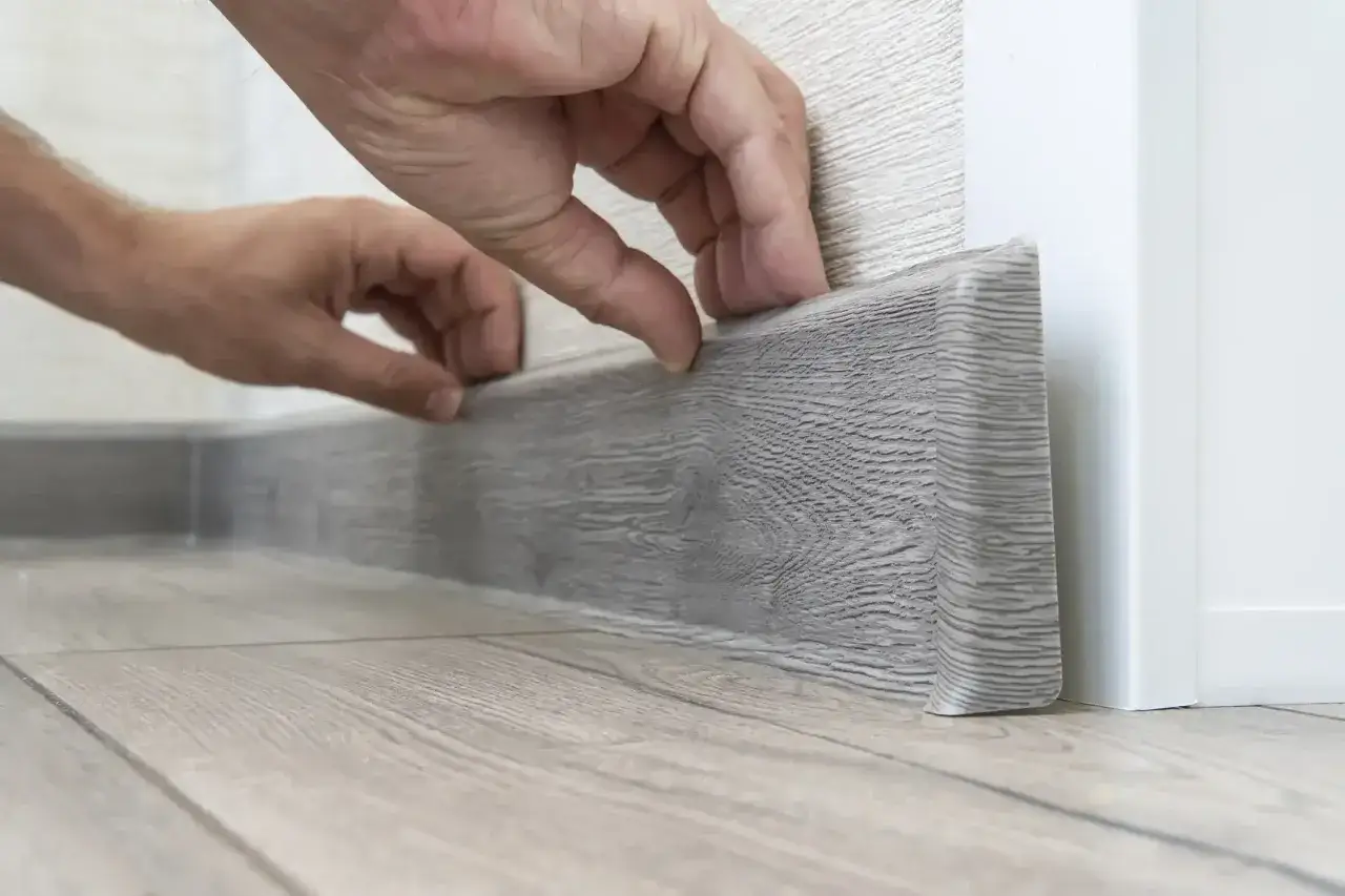 Hände montieren eine graue Sockelleiste an einem Laminatboden. Die Kosten für das Laminat verlegen sind oft überschaubar.