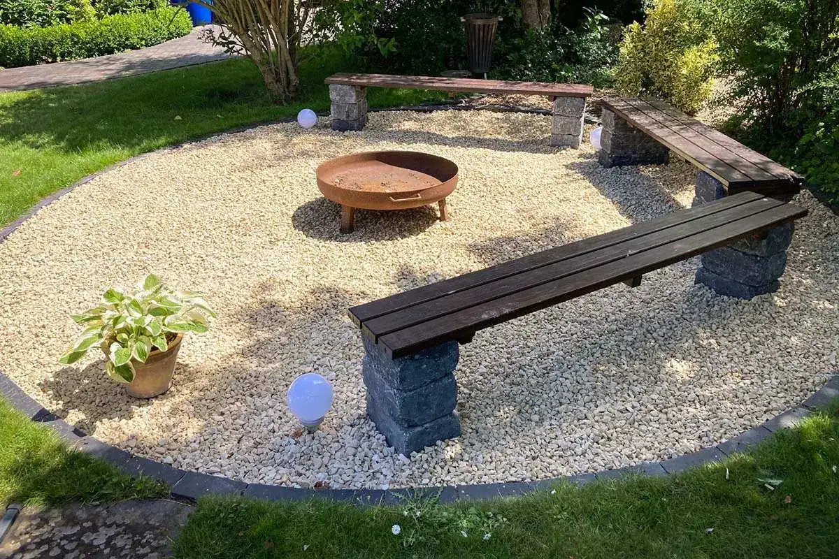 Gemütliche Feuerstelle auf einer Kies Terrasse mit Bänken und Beleuchtung.