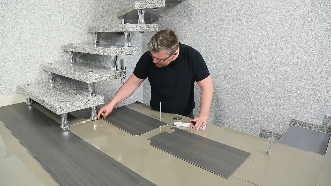 Mann verlegt PVC-Boden. Die grauen Planken werden präzise zugeschnitten und neben einer modernen Steintreppe platziert.