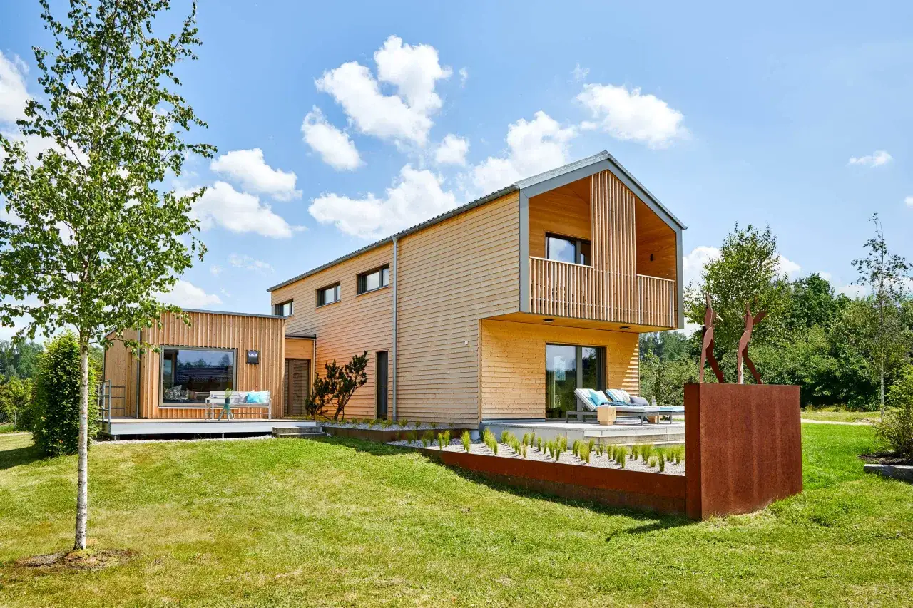 Modernes Fertighaus mit Holzfassade auf einem schmales Grundstück, mit Terrasse, Liegestühlen und Skulptur im Garten.
