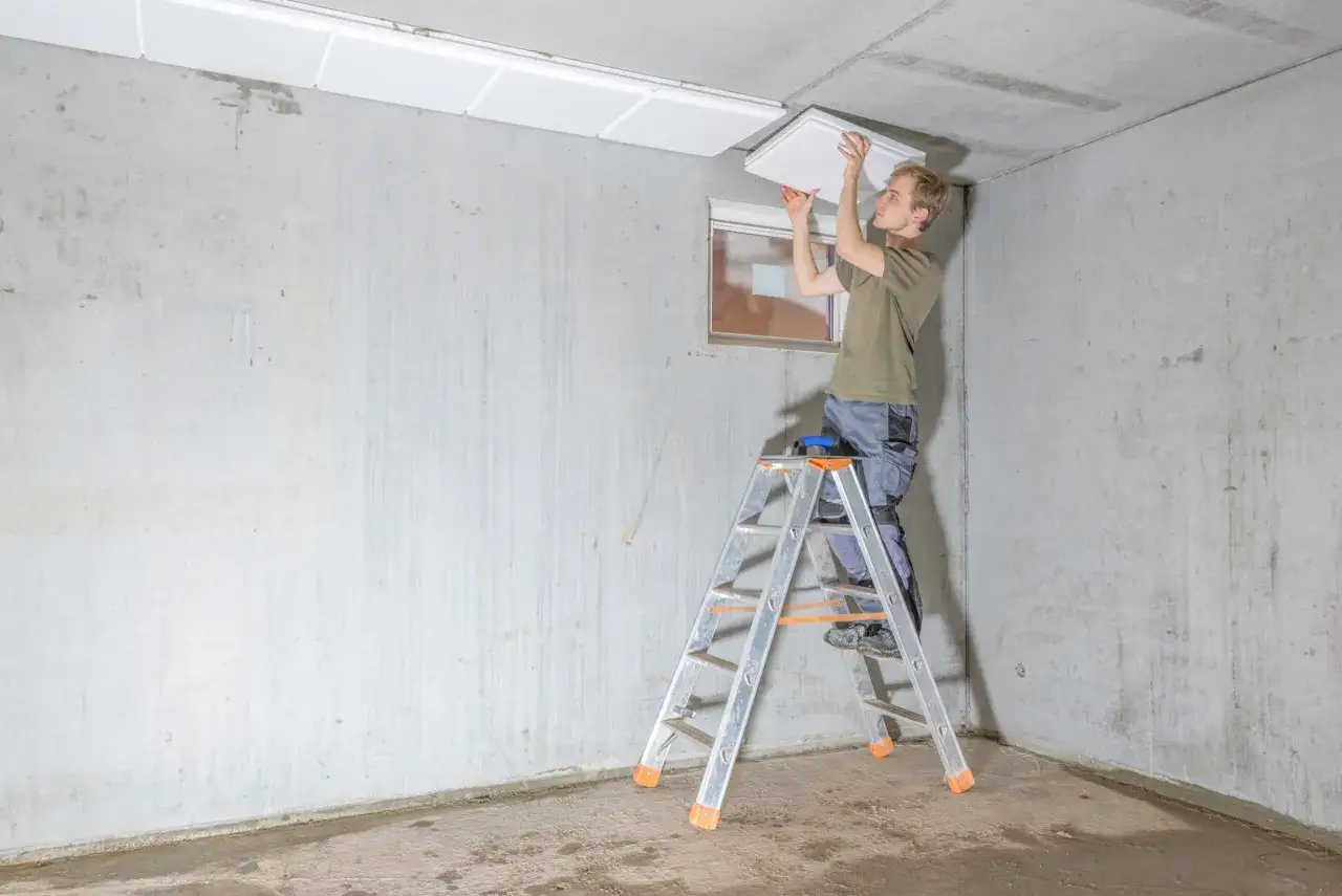 Mann auf Leiter montiert weiße Platten an Beton-Kellerdecke zur Isolierung.