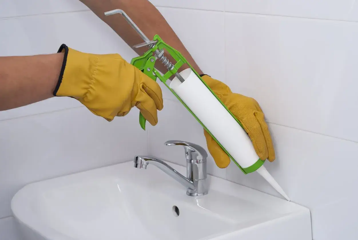 H&auml;nde mit gelben Handschuhen f&uuml;hren eine Kartuschenpresse, um Silikonfugen am Waschbecken zu erneuern.