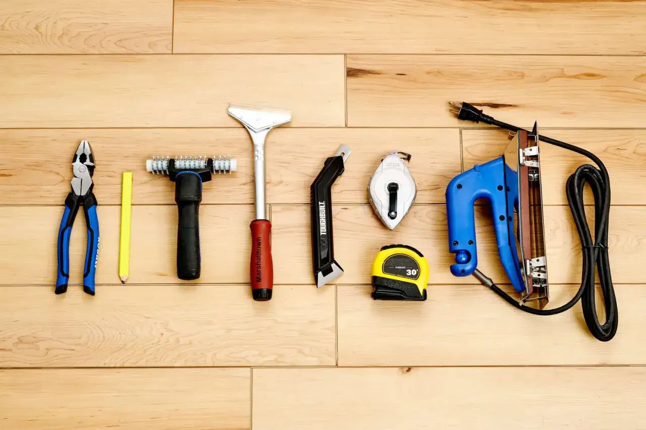 Werkzeuge f&uuml;r das Teppich verlegen: Zange, Bleistift, Teppichmesser, Ma&szlig;band, Kreideleine und ein elektrischer Tacker liegen auf Holzdielen.