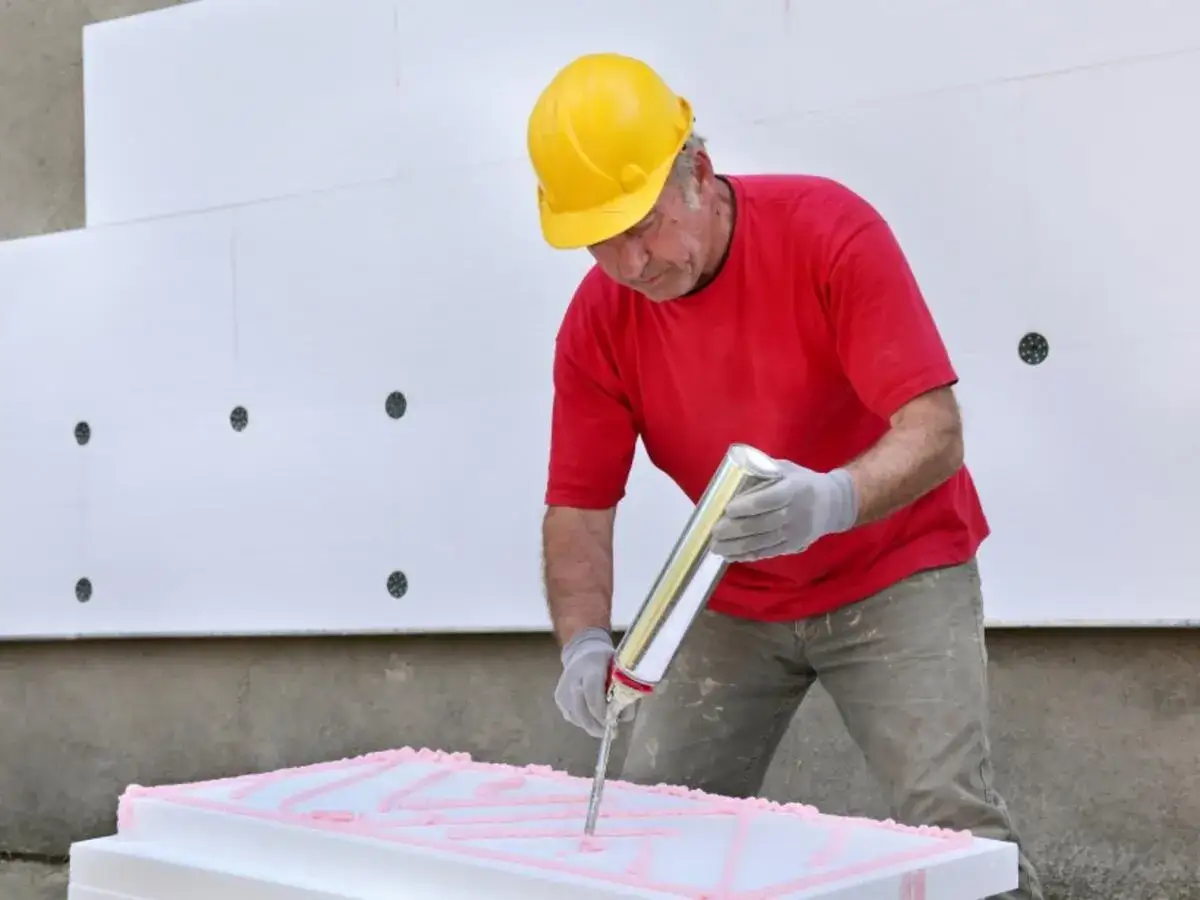 Mann mit gelbem Helm und rotem T-Shirt tr&auml;gt Kleber auf Styrodur-Platten auf, um sie zu isolieren.