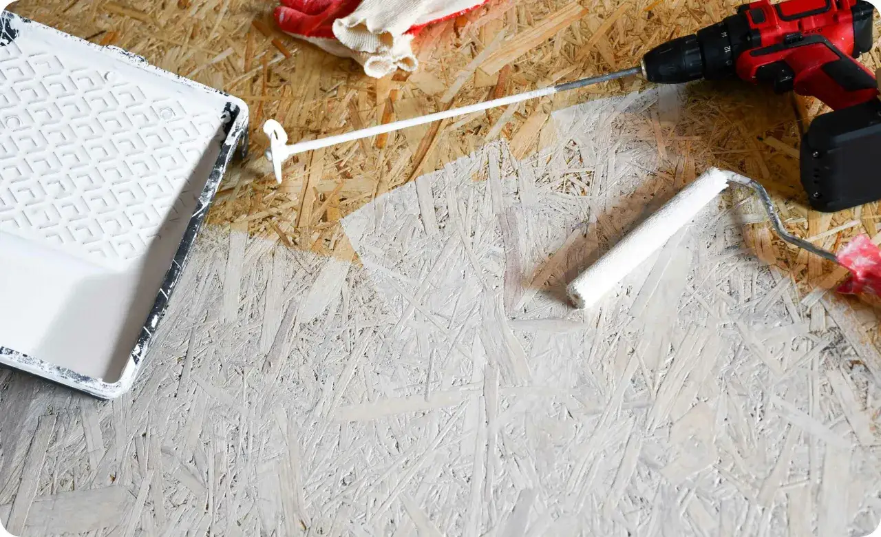Beim Streichen von OSB-Platten mit weißer Farbe: Ein Farbroller und eine Farbwanne liegen bereit, daneben eine Bohrmaschine und Handschuhe.