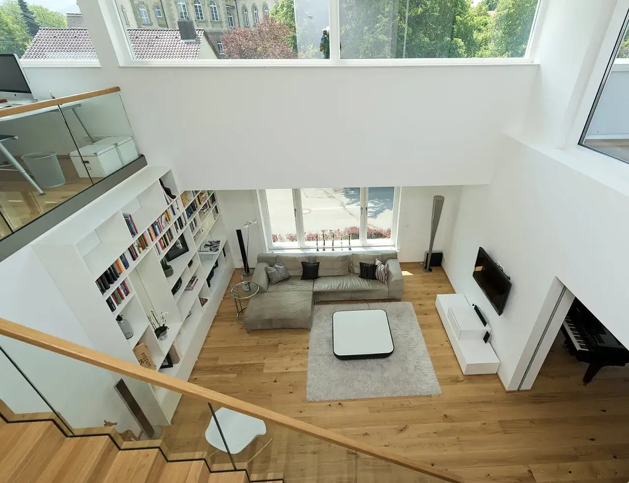 Ein offener Wohnbereich mit viel Luftraum und moderner Architektur. Ein großes Sofa, Bücherregale und ein Klavier prägen den Raum.