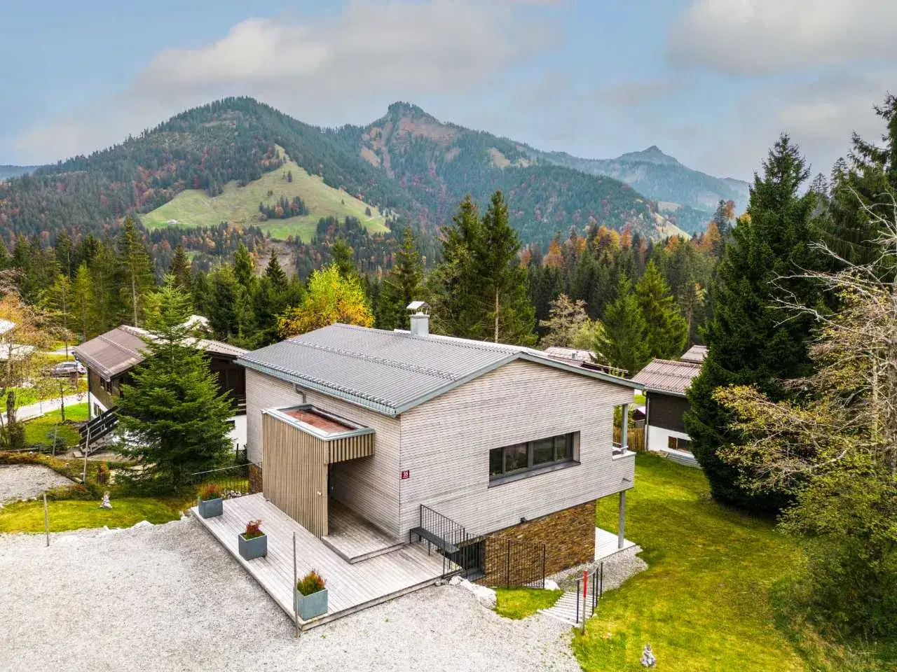 Modernes Haus in Hanglage mit Holzfassade, umgeben von B&auml;umen und Bergen im Herbst.