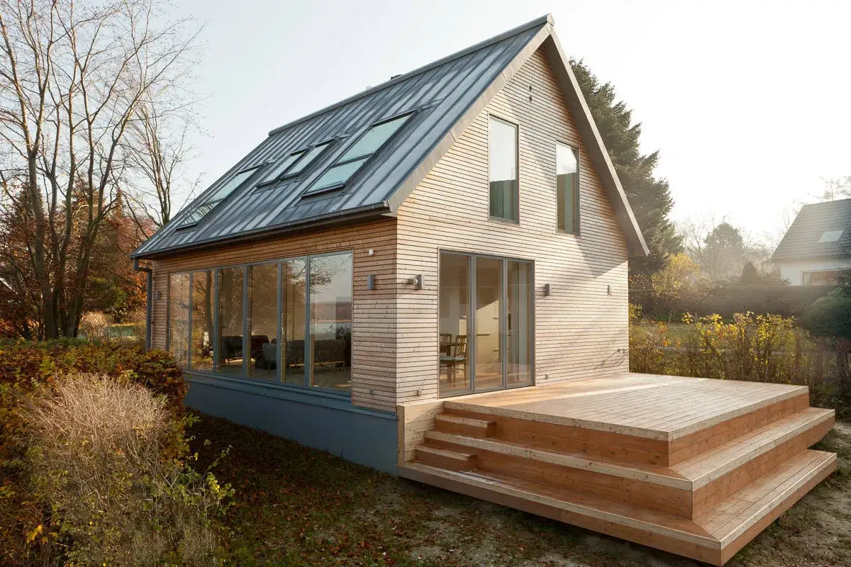 Modernes Holzhaus mit großen Fenstern und Dachgauben. Eine Holzterrasse mit Stufen lädt zum Verweilen ein. Ein Beispiel, wie man ein low budget haus bauen kann.