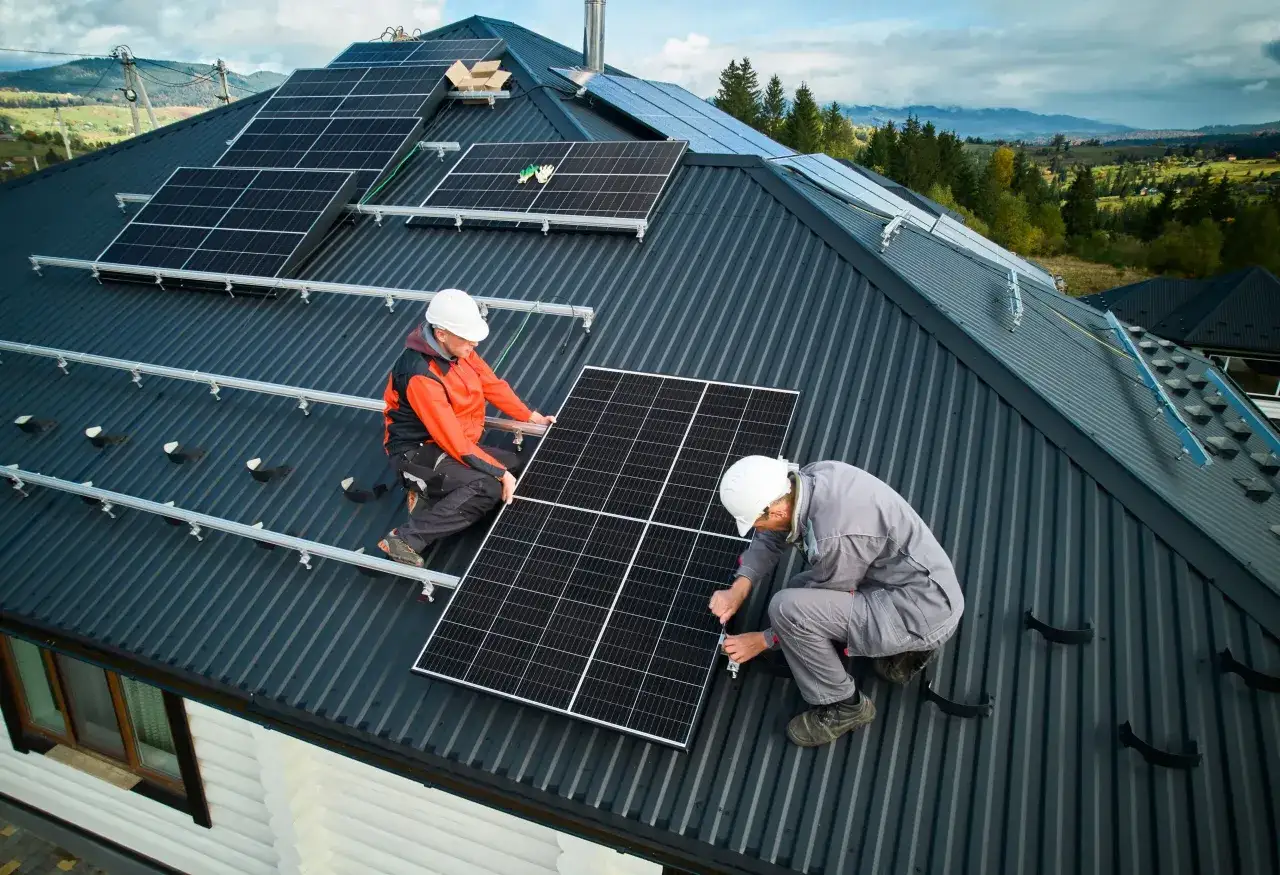 Zwei Handwerker installieren PV-Module auf einem dunklen Metalldach. Die Sonne scheint auf die Paneele, während die Landschaft im Hintergrund grün ist.