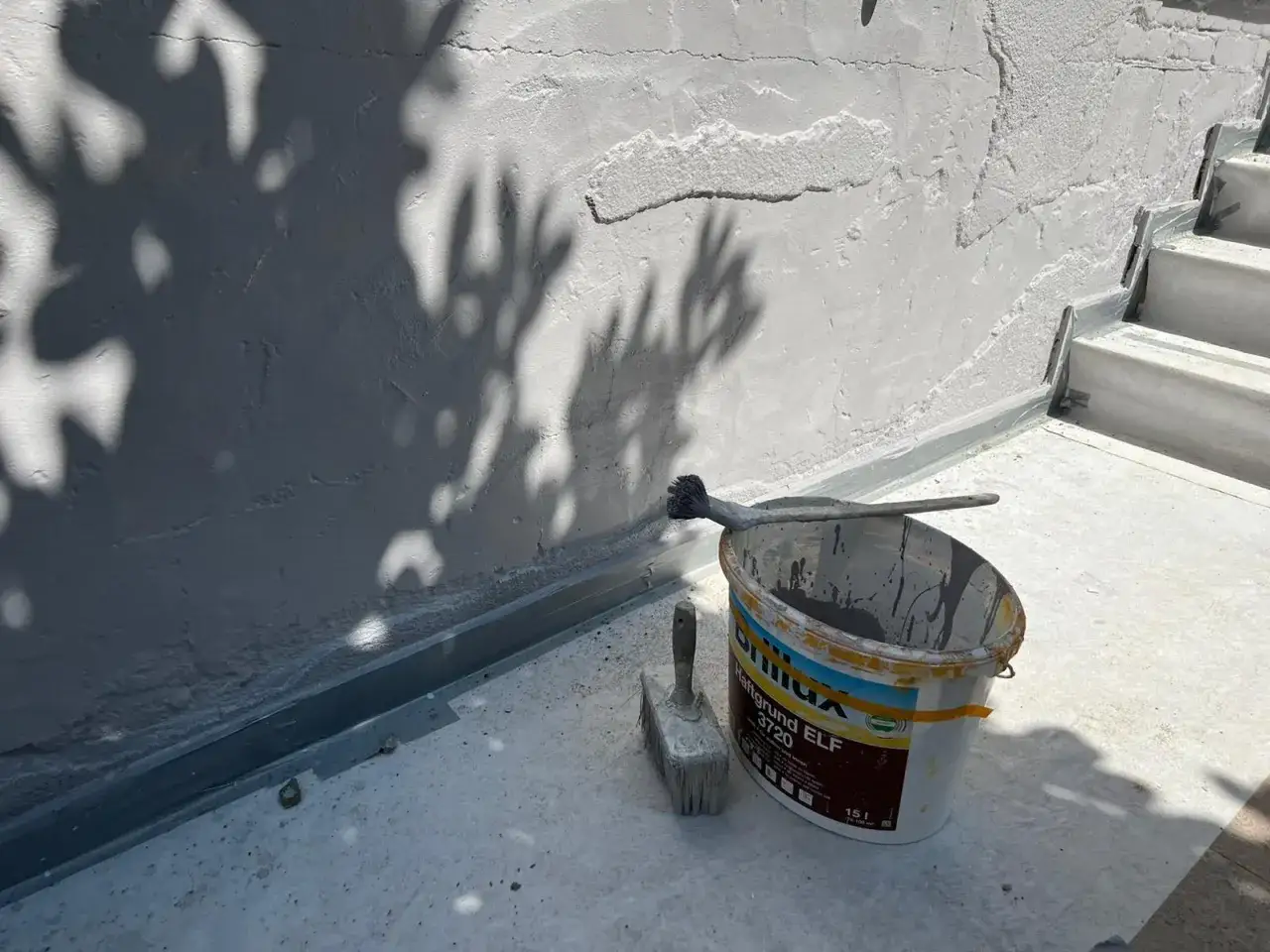 Werkzeug und Farbeimer bereit zum Sockelputz ausbessern. Schatten von Bl&auml;ttern tanzen auf der Wand.
