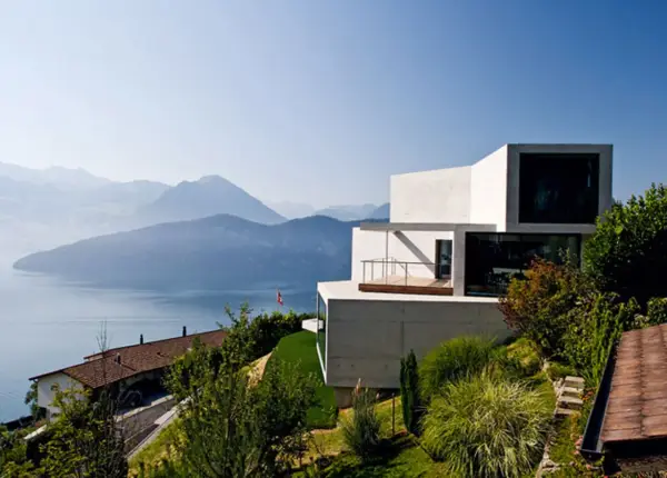 Modernes Haus in Hanglage mit Blick auf einen See und Berge. Ein traditionelles Haus liegt tiefer am Hang.