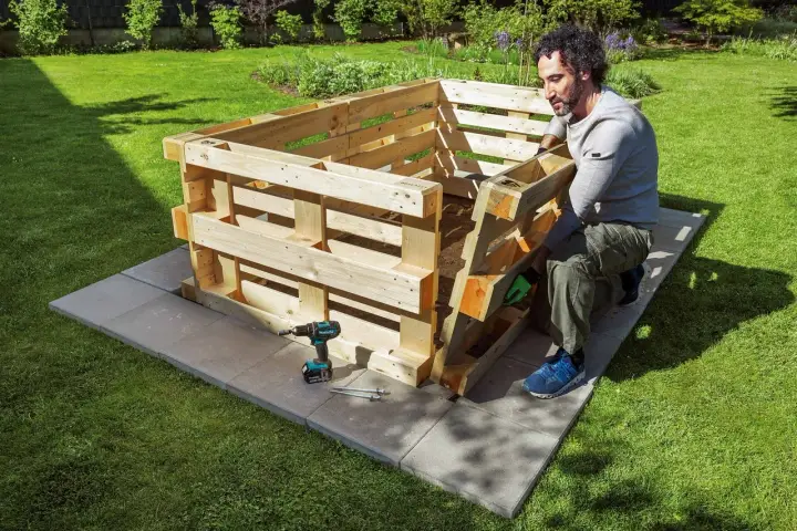 Mann baut ein **Hochbeet aus Paletten** im Garten. Werkzeug und Material liegen bereit.