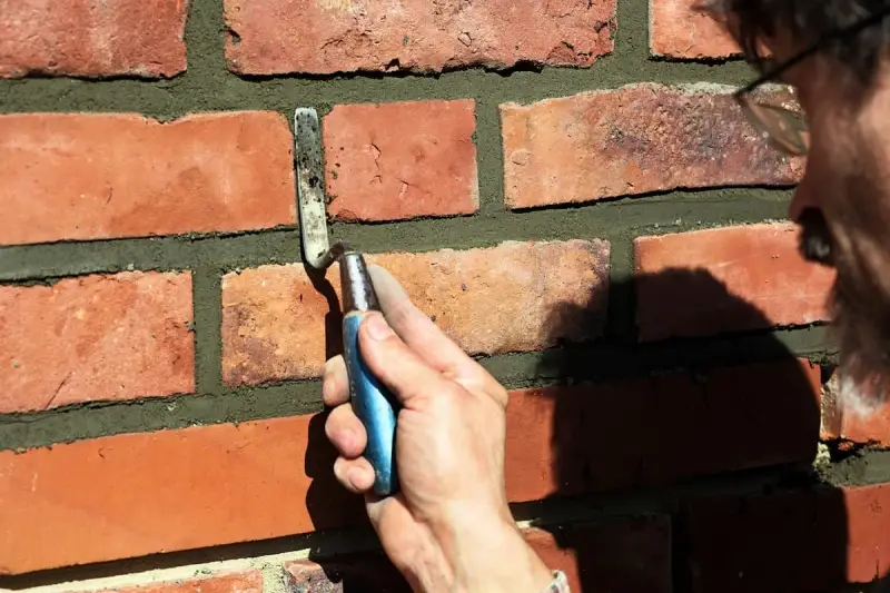 Hand mit Kelle bessert Zementfugen einer Ziegelwand aus.